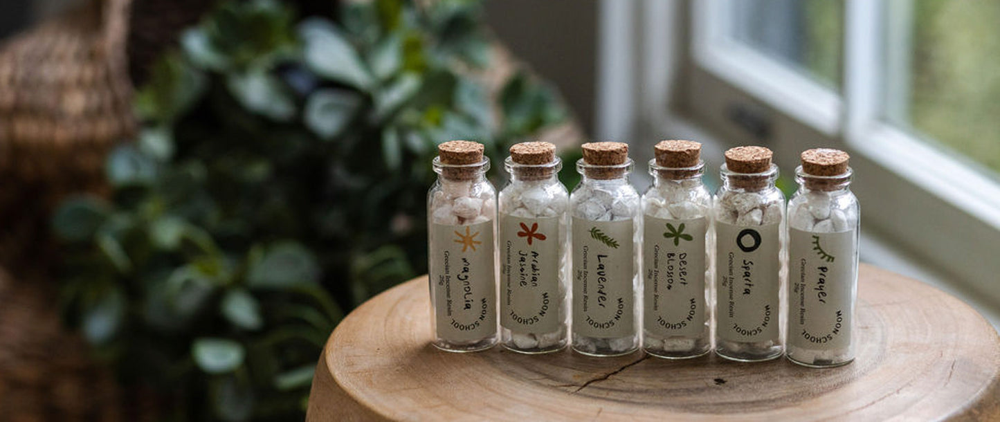 Six bottles of Grecian incense resins arranged on a wooden table, each labeled with its botanical scent blend. Natural ritual tools by Moon School.