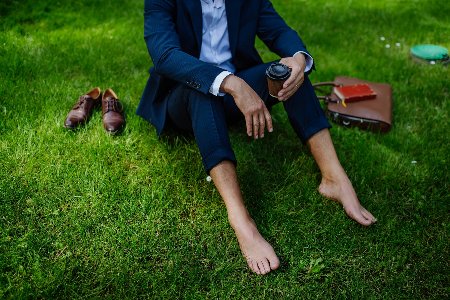 Professional in suit walking barefoot on grass, holding coffee—a moment of midday reset and calm.