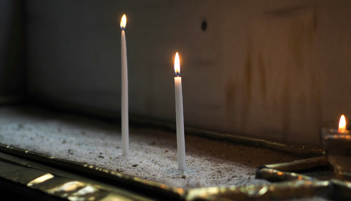 Two thin white candles lit in sand, warm golden light, a third candle softly out of focus in the background