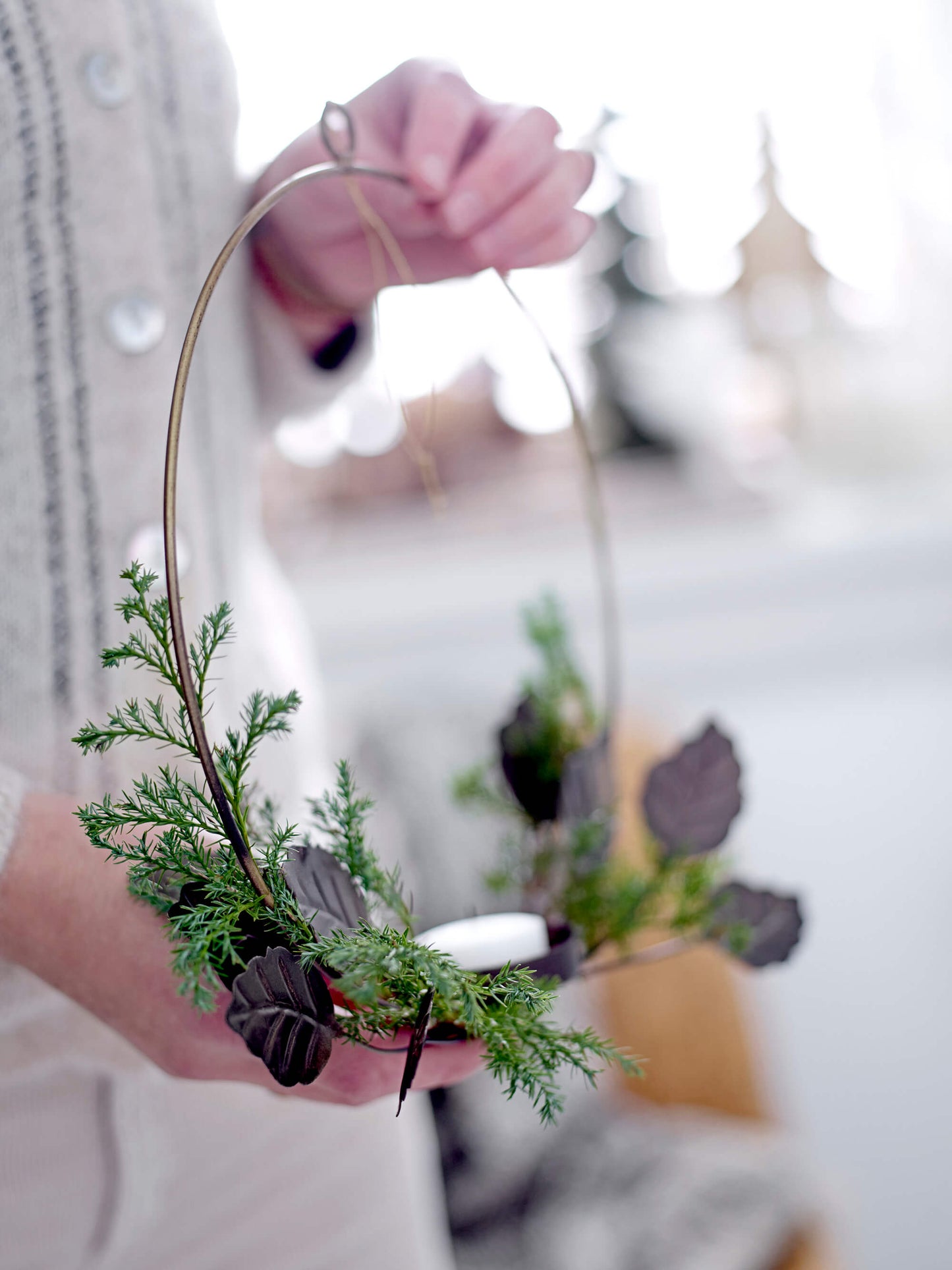A lady holding the Ivalou Wall Votive by Bloomingville in her hand. The round design, adorned with brownish metal leaves, features added decor of fern leaves elegantly wrapped around it. Measuring 28cm in diameter and 6cm in width, it provides a warm and cozy light, suitable for any room. Note: For safety, do not leave lit candles unattended or in a draft. Enhance its look by adding your own beautiful decorations.