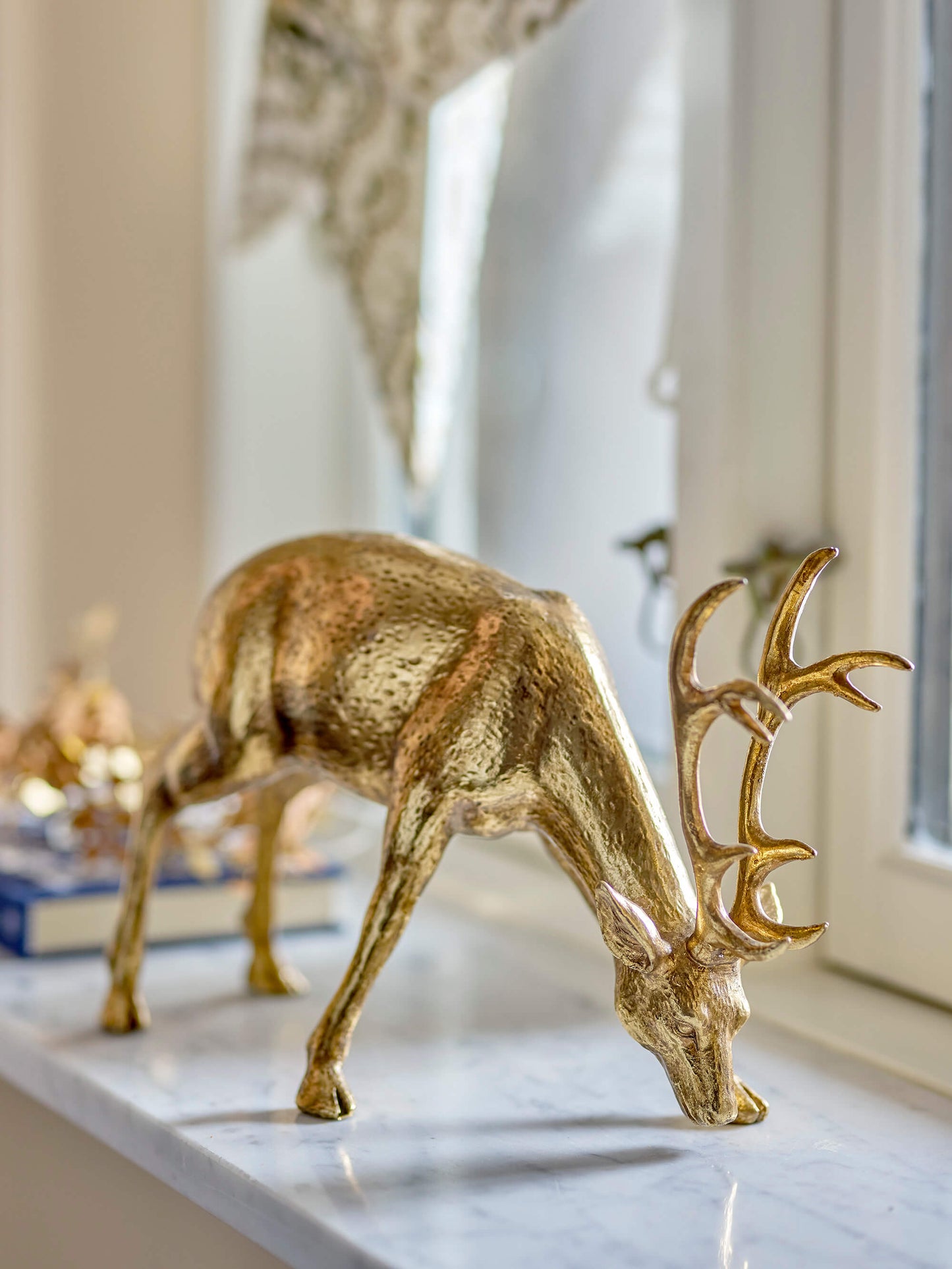 Keera Deer by Bloomingville elegantly displayed on a window sill shelf. The golden polyresin deer adds a festive touch to any space. Size: 19cm(H) x 33cm(L) x 13cm(W). Style it with spruce, pinecones, candles, or crystals for a natural and enchanting look.