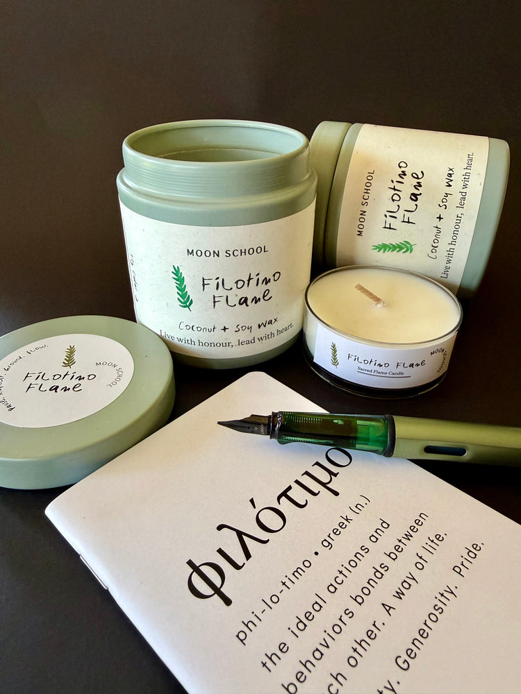 
                  
                    Two candles labeled 'Moon School Filotimo Flame' on a surface with a book and pen.
                  
                