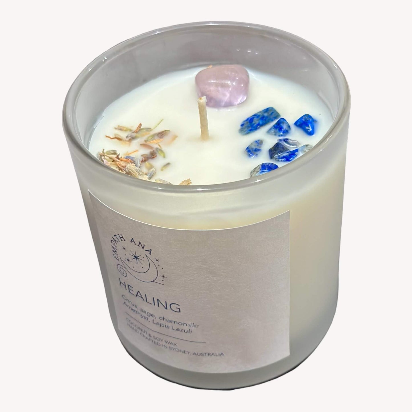 Empath Ana's Healing Intention Crystal Candle - Top view of the same candle jar, revealing Amethyst and Lapis Lazuli crystals embedded in the coconut/soy wax, surrounded by healing herbs. The crystals are complemented by an uplifting Citrus Sage & Chamomile fragrance